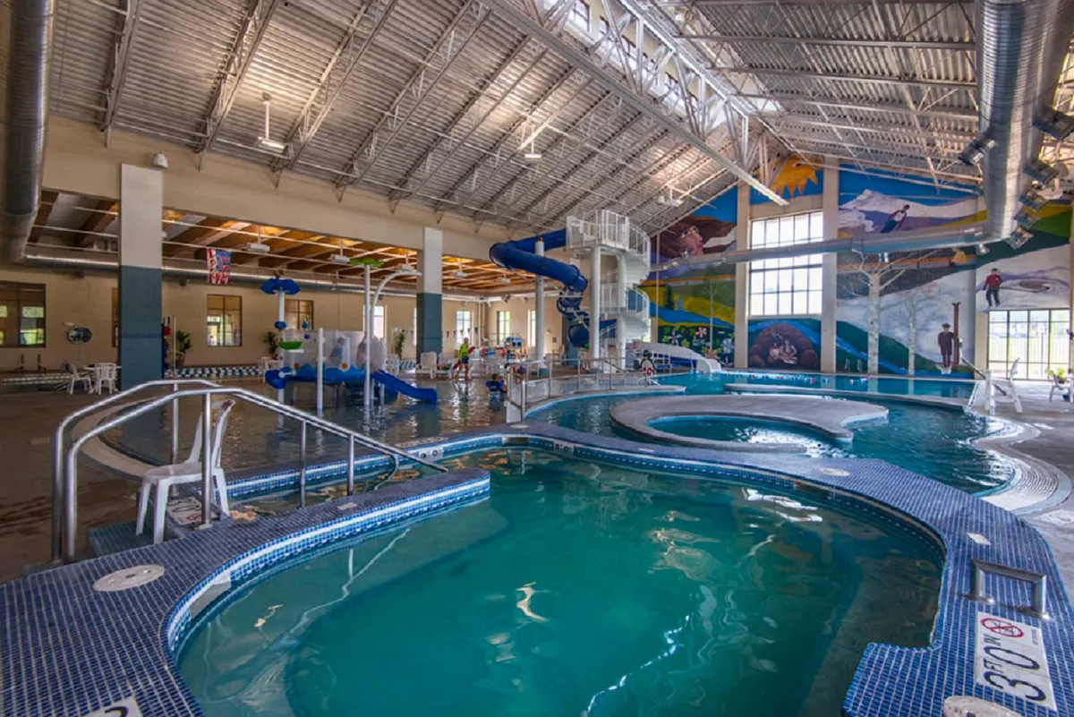 An energetic indoor aquatic center featuring colorful water slides, a swimming pool, and a mural on the wall. Families and children are enjoying water play and gathering around the pool area, creating a sense of lively, joyful activity.