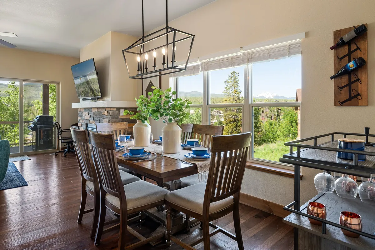 A warm, inviting dining area with a spacious table positioned near large windows, allowing for natural light and views of the outdoors. Decorative plants and an elegant lighting fixture add to the cozy, family-friendly ambiance.
