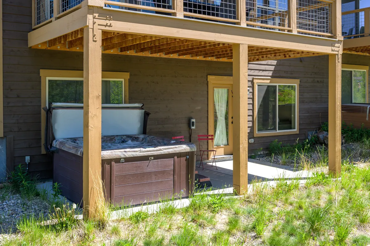 A welcoming patio area featuring a private hot tub, wooden support beams, and a side entrance door, surrounded by natural landscaping. The area is designed for relaxation with comfortable seating, perfect for socializing or unwinding after outdoor adventures.