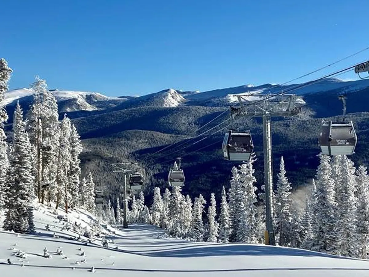 A panoramic view of ski lift gondolas gliding over snow-covered slopes and distant mountains. Pine trees frame the scene, emphasizing the resort’s alpine setting and the thrill of winter sports.