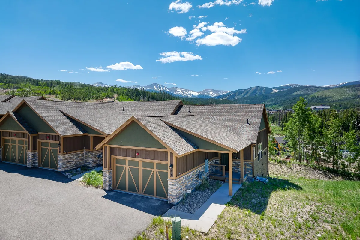 The image showcases a stunning mountain chalet with modern architectural details, nestled against a breathtaking backdrop of the Rocky Mountains. The driveway curves invitingly toward the home, while the spacious porch and yard suggest opportunities for outdoor gatherings, sunset watching, and soaking in the peaceful alpine atmosphere.
