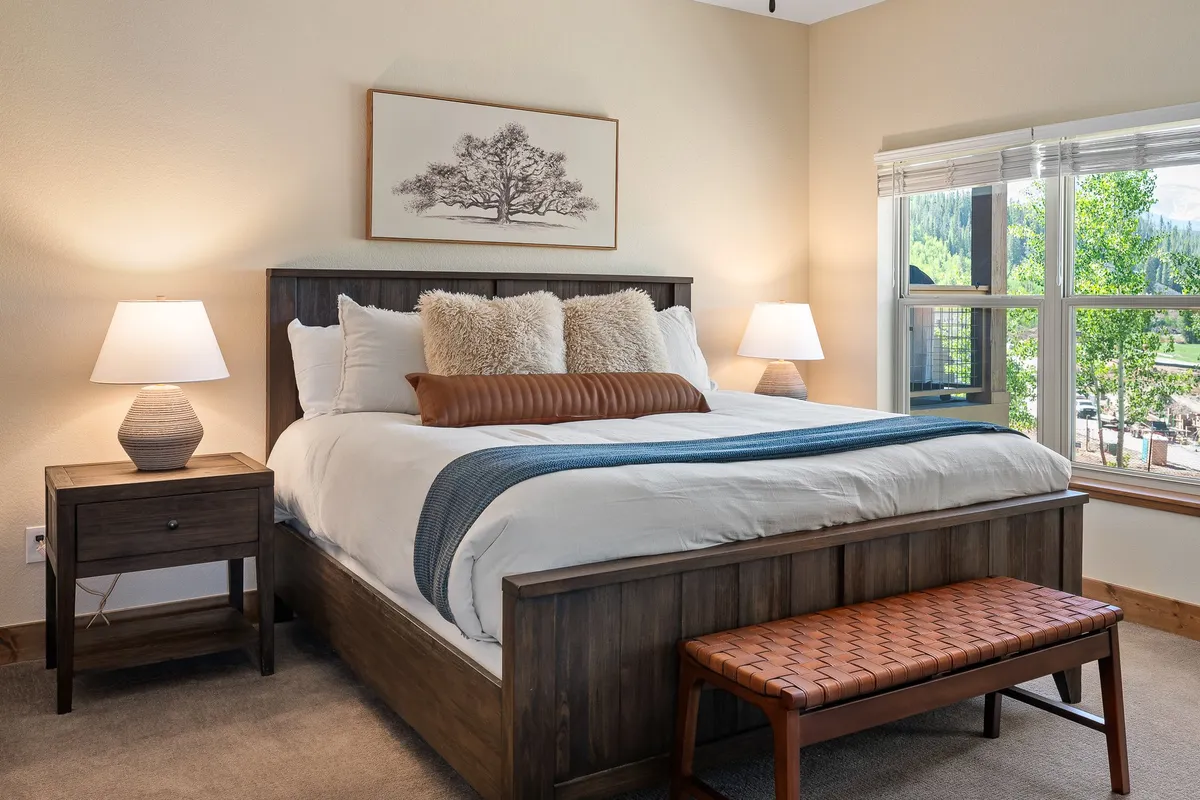 A serene bedroom with a comfortable bed, nightstands, decorative artwork, and sunlight streaming through a window with a view. The styling suggests luxury and tranquility, with premium linens and a calming color palette that fits the description of spa-inspired, private retreats.