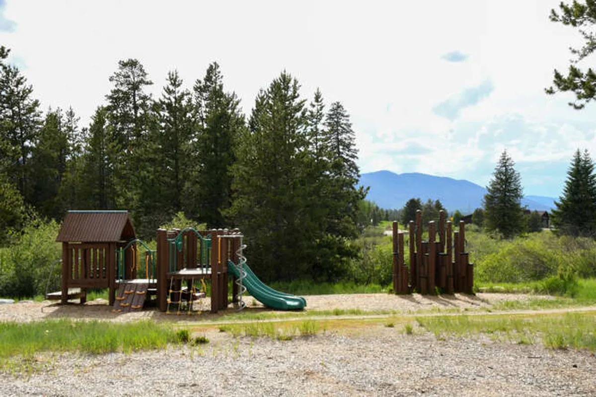 A bright and welcoming playground area featuring a slide, climbing frame, and surrounded by trees, with the mountains visible in the background. Children are actively playing on the equipment while families gather and picnic nearby, creating a lively, joyful scene.