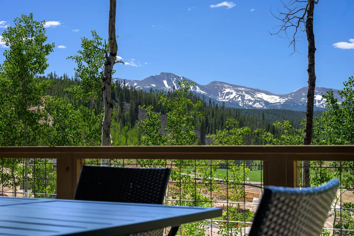 An outdoor seating area overlooks a stunning Rocky Mountain range, framed by green foliage. The space is ideal for relaxing, dining, or simply soaking up the breathtaking mountain scenery, particularly at sunset.