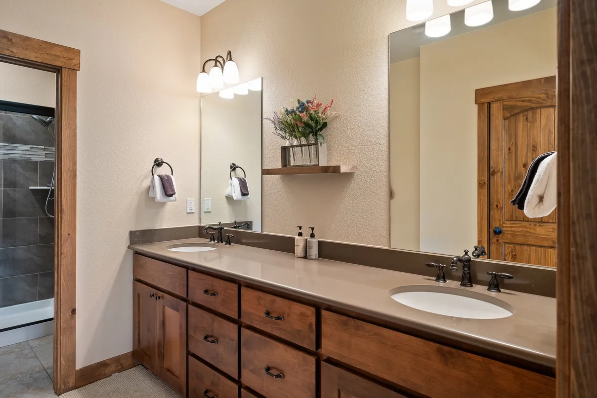 A contemporary bathroom featuring a double sink vanity, a large mirror, wooden cabinetry, and a decorative shelf with fresh flowers. The overall feel is calm and spa-like, ideal for self-care or family routines before new adventures.