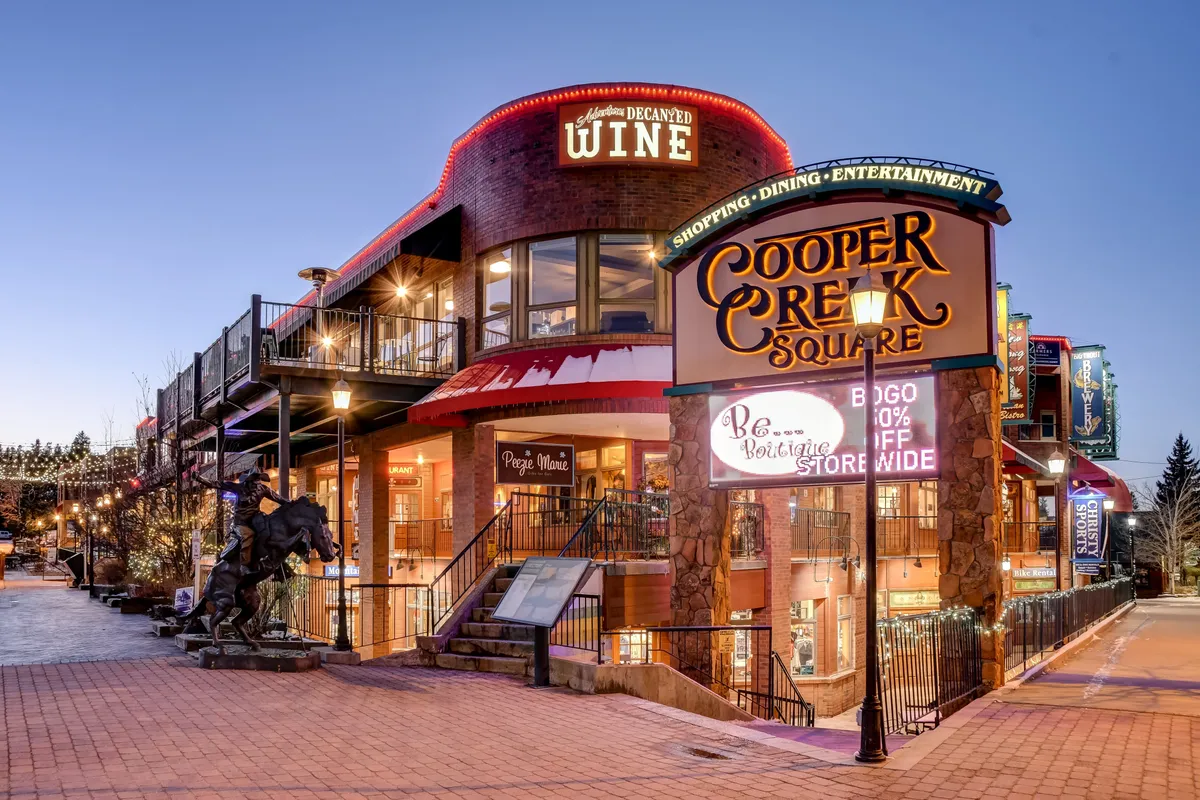 A lively snapshot of Cooper Creek Square, featuring vibrant decorative lighting, people engaging in shopping and dining, and a prominent wine shop entrance. The energy of the area is palpable, with signs of seasonal festivals and a welcoming atmosphere for entertainment and socializing.