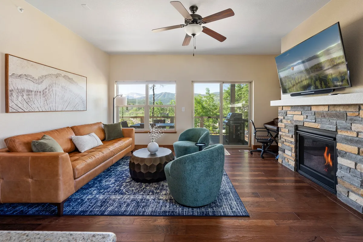 A cozy open-plan living area featuring a leather sofa, round coffee table, fireplace, large TV, and expansive windows that reveal stunning mountain vistas. The room is bathed in natural light, with inviting seating arranged to encourage relaxation, conversation, and group activities.